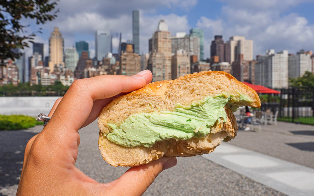 Dana holding a bagel with avocado cream cheese in front of the NYC skyline